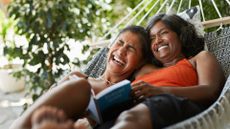 Woman laughing and lying down with daughter in hammock, reading a book on travel