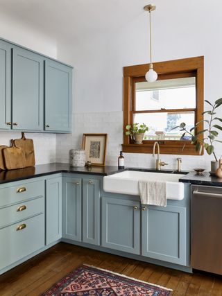 pale blue kitchen cabinetry with white walls, textured white backsplash tiles, wooden window to porch, hardwood floor, antique rug, large Butler sink, black granite countertop, brass faucet