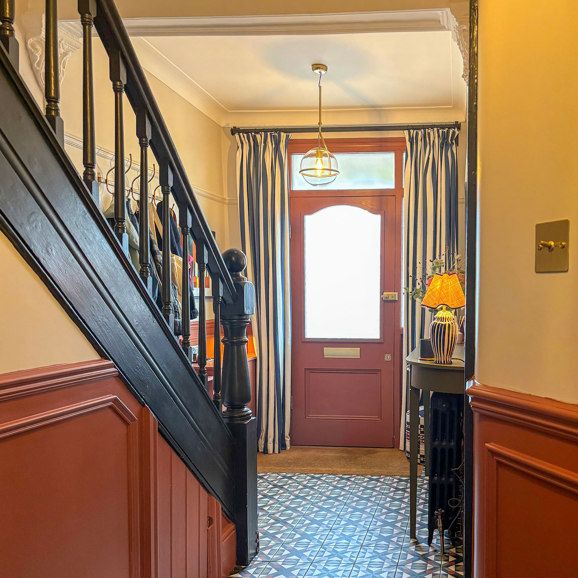 Entrance hallway in home showing inteior of terracotta painted door, double stripy door curtains, patterned tile floor, black painted banisters and walls with lower wall panelling