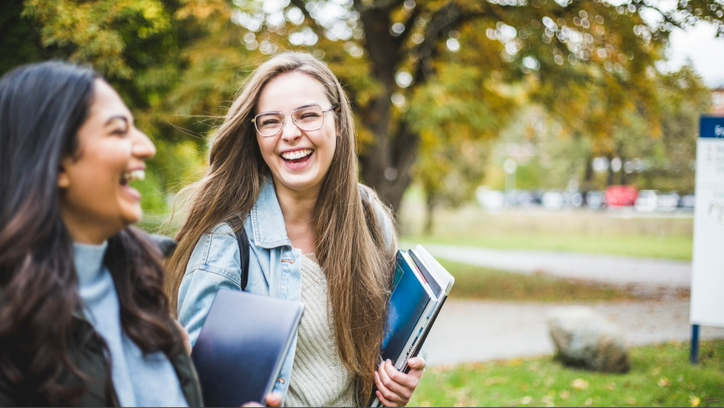 Two female students carrying files as they walk through green park laughing