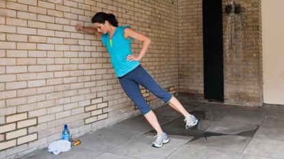 A woman performs a side plank against a wall. She is outside, with her right side facing the wall and her right elbow and forearm placed on the wall, at shoulder height. Her feet are stepped away from the wall. 