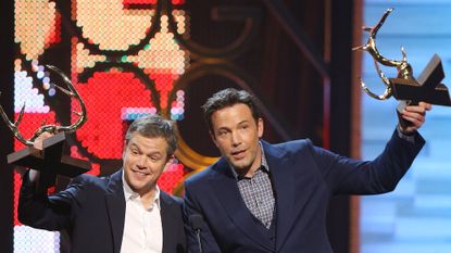 Matt Damon and Ben Affleck on stage together raising their award trophies in celebration.