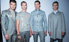 Four models wearing Brioni clothing, two with grey suit jackets with gold and turquoise accent, one with grey coat and one with grey golf shirt. 