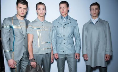 Four models wearing Brioni clothing, two with grey suit jackets with gold and turquoise accent, one with grey coat and one with grey golf shirt. 