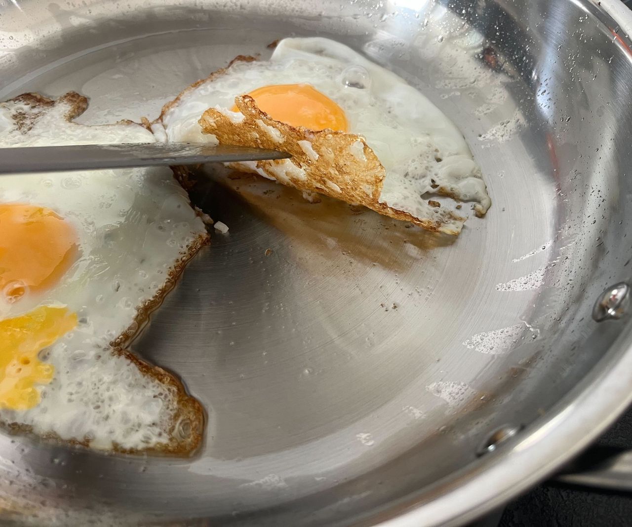 A fried egg with a crispy base being lifted by a spatula in a stainless teel frying pan.
