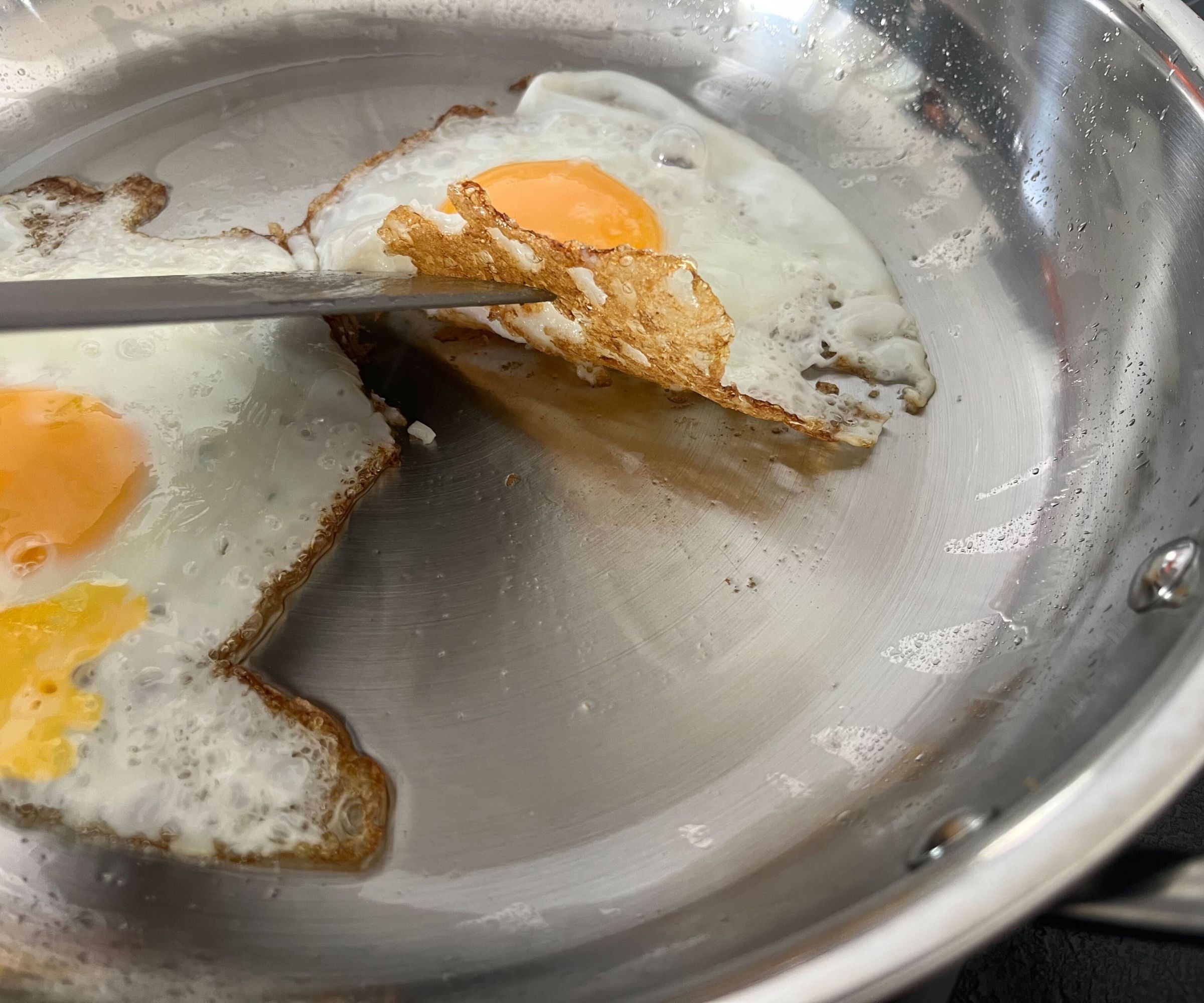 A fried egg with a crispy base being lifted by a spatula in a stainless teel frying pan.