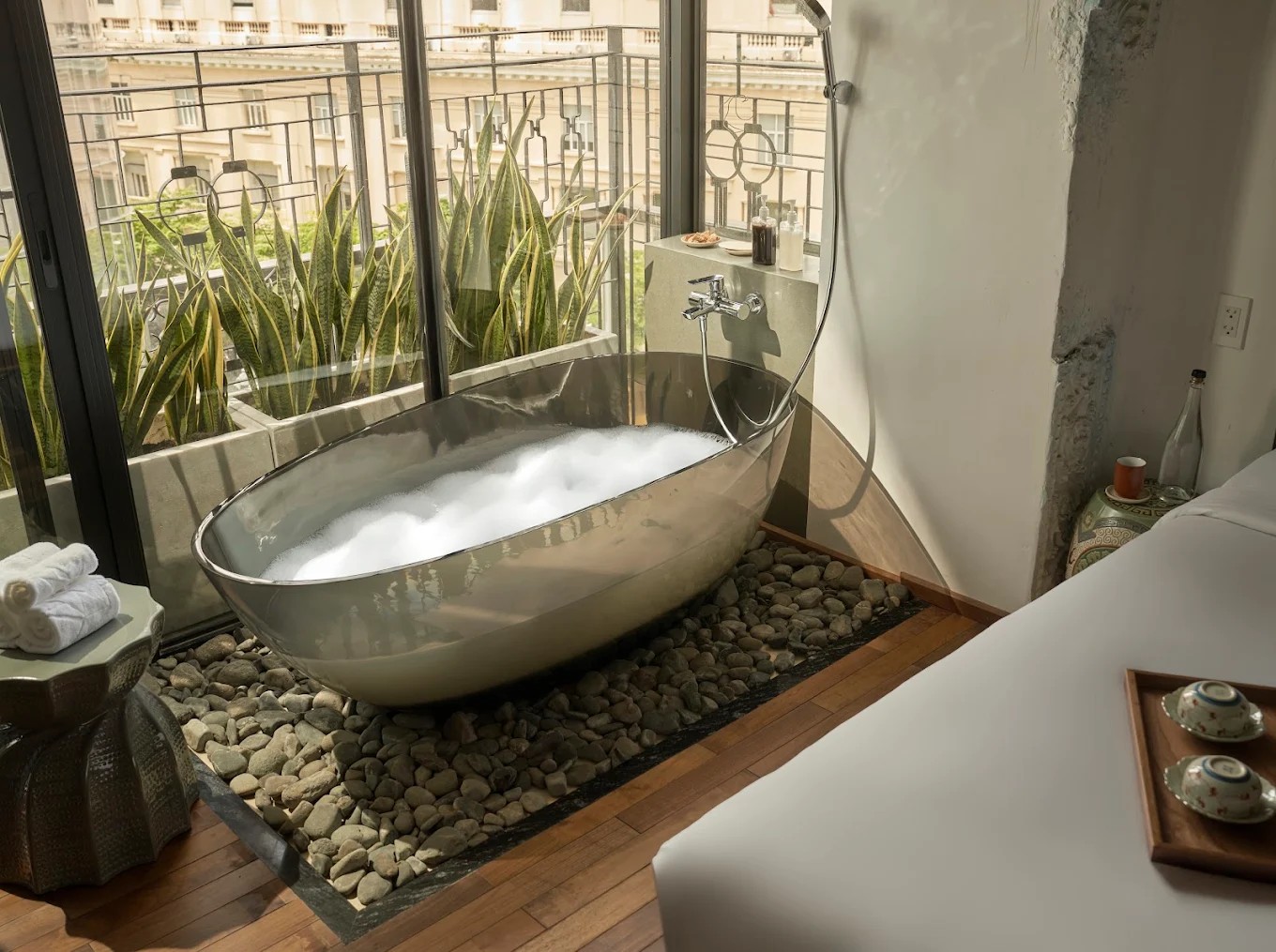 An in-suite standing bathtub in chrome, styled atop a bed of rocks, next to trailing plants and a neutral interior design scheme of woods, ceramics, and white textiles.