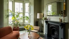 A living room painted in a green color with bay windows and beneath the windows is a small radiator also painted in green. In the living room there is also a fireplace, sofa and coffee table with a vase and flowers on top