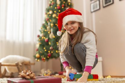 Happy little girl wearing Santa hat sitting on the floor by nicely decorated Christmas tree having fun playing with toy blocks on Christmas day