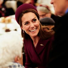 Kate Middleton wears a wine red coat dress and matching hat and laughs