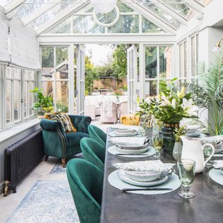 a glass conservatory willed with plants with a large dining table and green upholstered velvet chairs