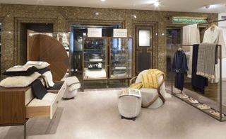A clothing store with a clothing on a rail on the right, a wooden chest of drawers on the left, glass display cabinets on the back wall and a chair and a foot stool in the middle of the room.
