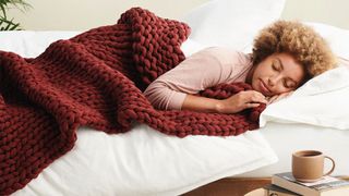 A woman lying on a bed, covered by a red weighted blanket, with a coffee cup and books by her bedside
