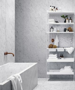 A white bathroom with bathtub, brass faucet and modular shelving and gray wall and floor tile