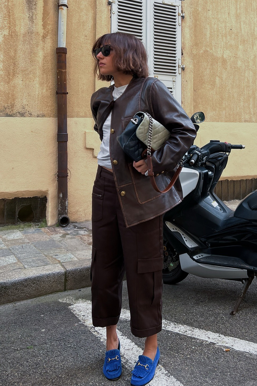A woman wearing a brown leather jacket, brown cargo pants, blue loafers, a brown and gray quilted handbag, and black sunglasses