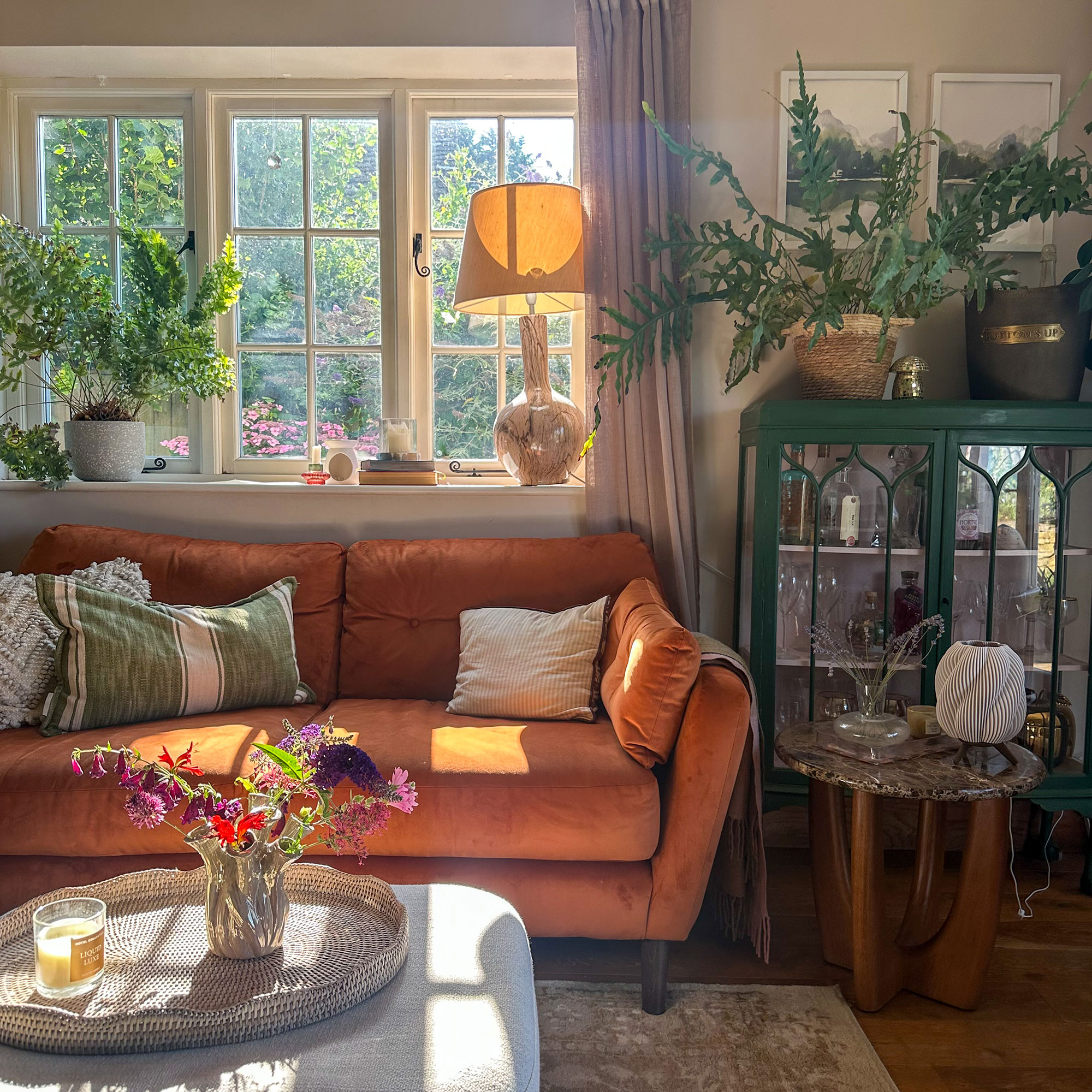 Living room with burnt orange sofa beneath windows, opposite ottoman with woven tray holding vase of flowers