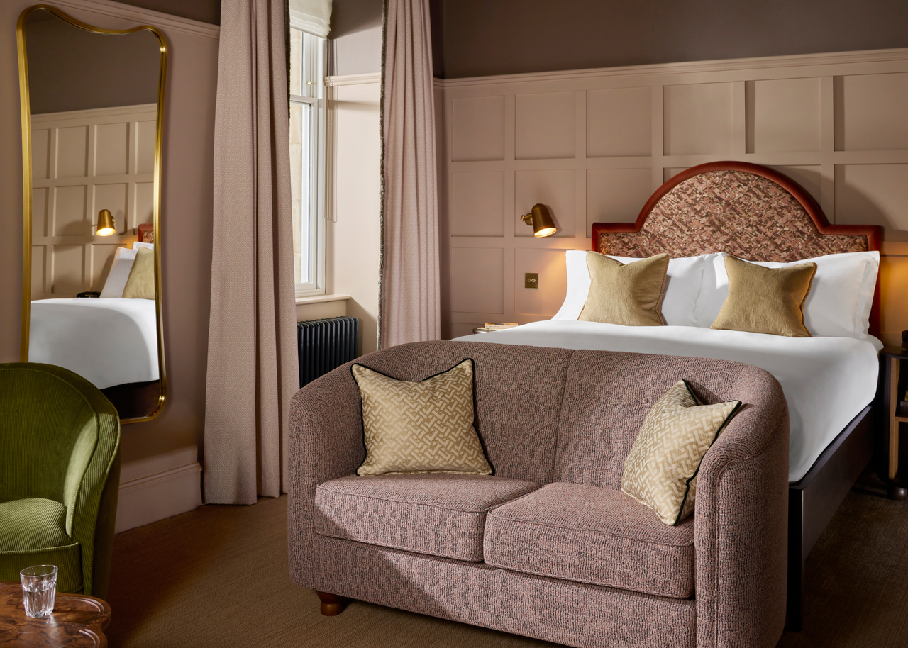 A beige bedroom with gray capped ceilings, a long mirror, a green armchair, a wood side table with a glass of water by a brown couch by the foot of a bed, and a bed with a statement headboard
