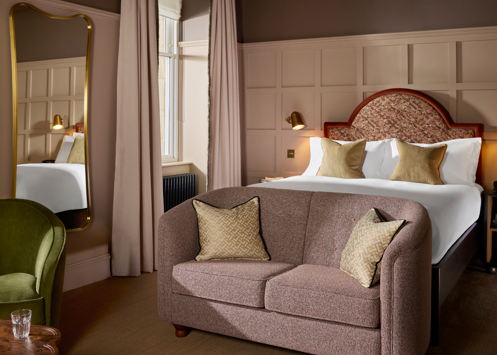 A beige bedroom with gray capped ceilings, a long mirror, a green armchair, a wood side table with a glass of water by a brown couch by the foot of a bed, and a bed with a statement headboard