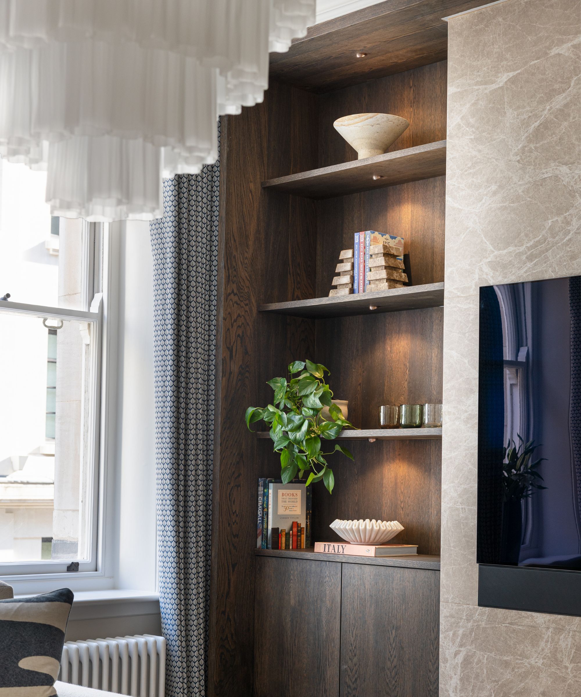 A built-in dark wood shelving unit is set into a recessed wall beside a large window dressed with patterned curtains. The shelves are illuminated by small, warm-toned spotlights, highlighting a mix of decorative items including books, a leafy houseplant, and small ornaments.