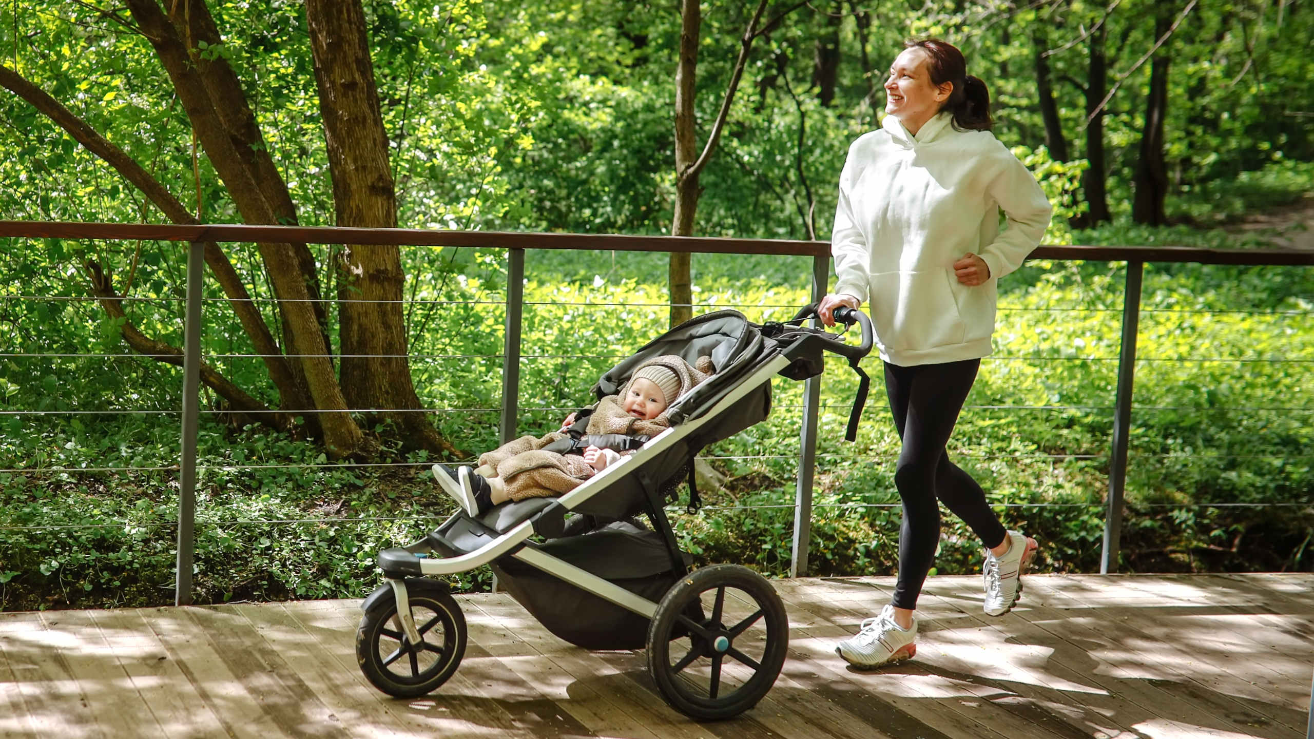 A woman runs along a wooden path pushing a baby in a three-wheeled stroller.