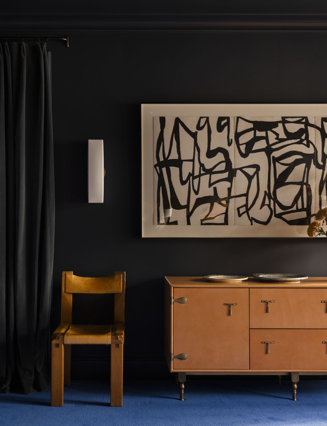 Image of a black room with an electric blue carpet. There is a warm, orange wood cabinet and matching accent chair, and a black and white abstract painting hanging over the cabinet.