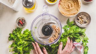 A food processor containing some nuts, on a worktop that also has fresh herbs, homemade peanut butter, oil and seeds in bowls