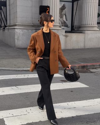 A woman wears a brown blazer, black top, black trousers, black bag, and black ankle boots.