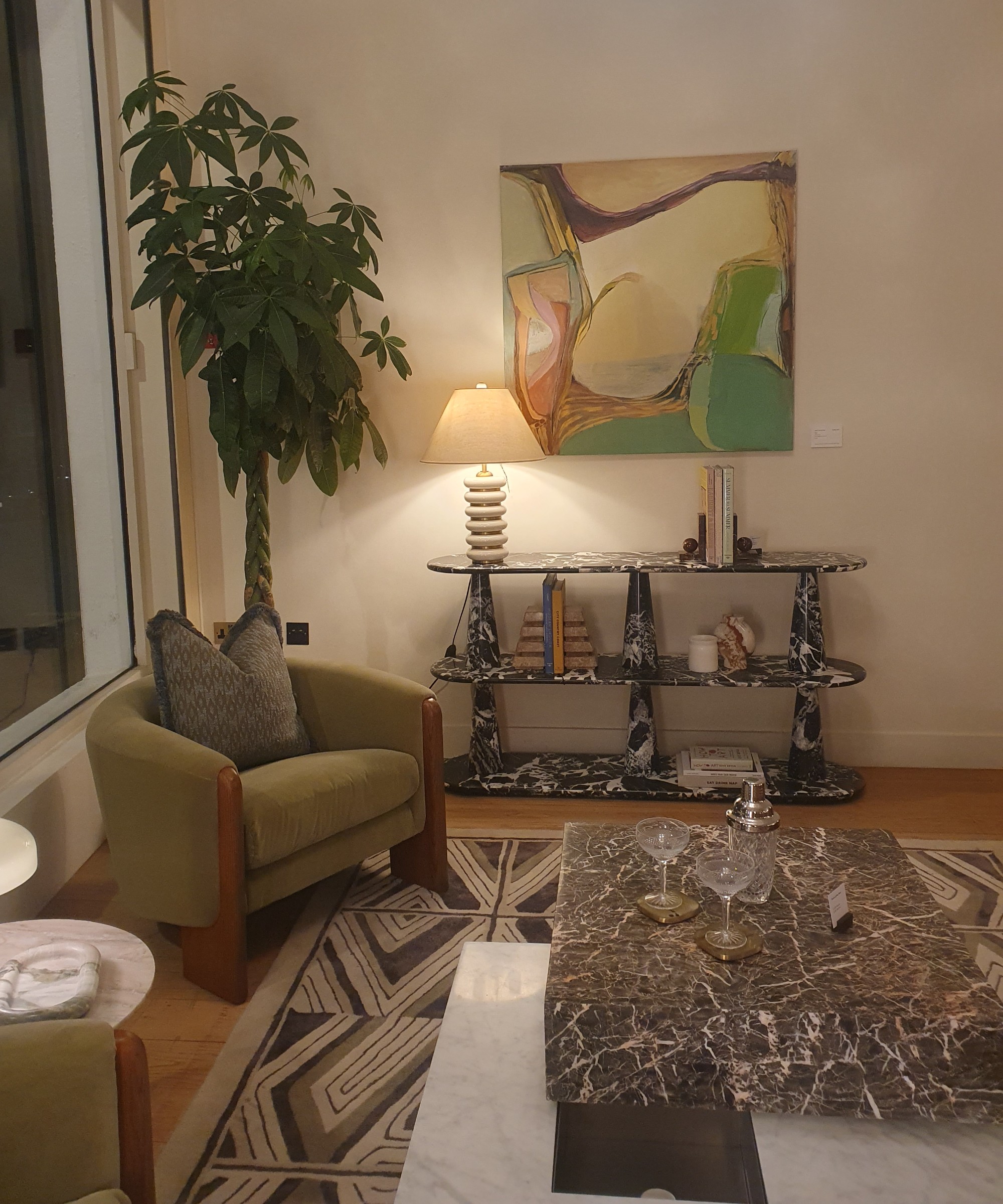 A section of the Soho Home Notting Hill store with a green velvet accent chair, a marble coffee table and console table and a switched on table lamp with a totem-style base