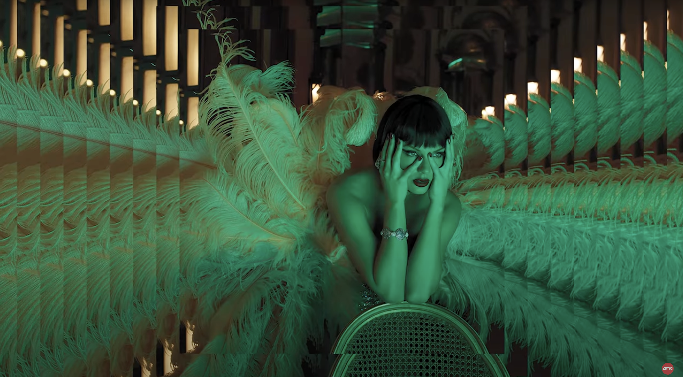 Taylor Swift, with dark hair and bangs, leans her arms on a chair, surrounded by large, dramatic green feathers in a darkly lit, glamorous setting.
