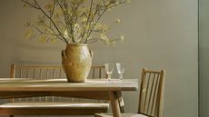 corner of modern dining room with earthy beige walls, wooden dining table and benched with vase of foliage
