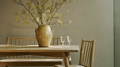 corner of modern dining room with earthy beige walls, wooden dining table and benched with vase of foliage