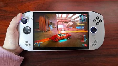 Hand holding Lenovo Legion Go S handheld on woodgrain desk with Cyberpunk 2077 gameplay on screen.