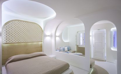 A hotel bedroom with a bed, white side table, brown corner sofa, patterned cushions, white walls and arched an doorway.