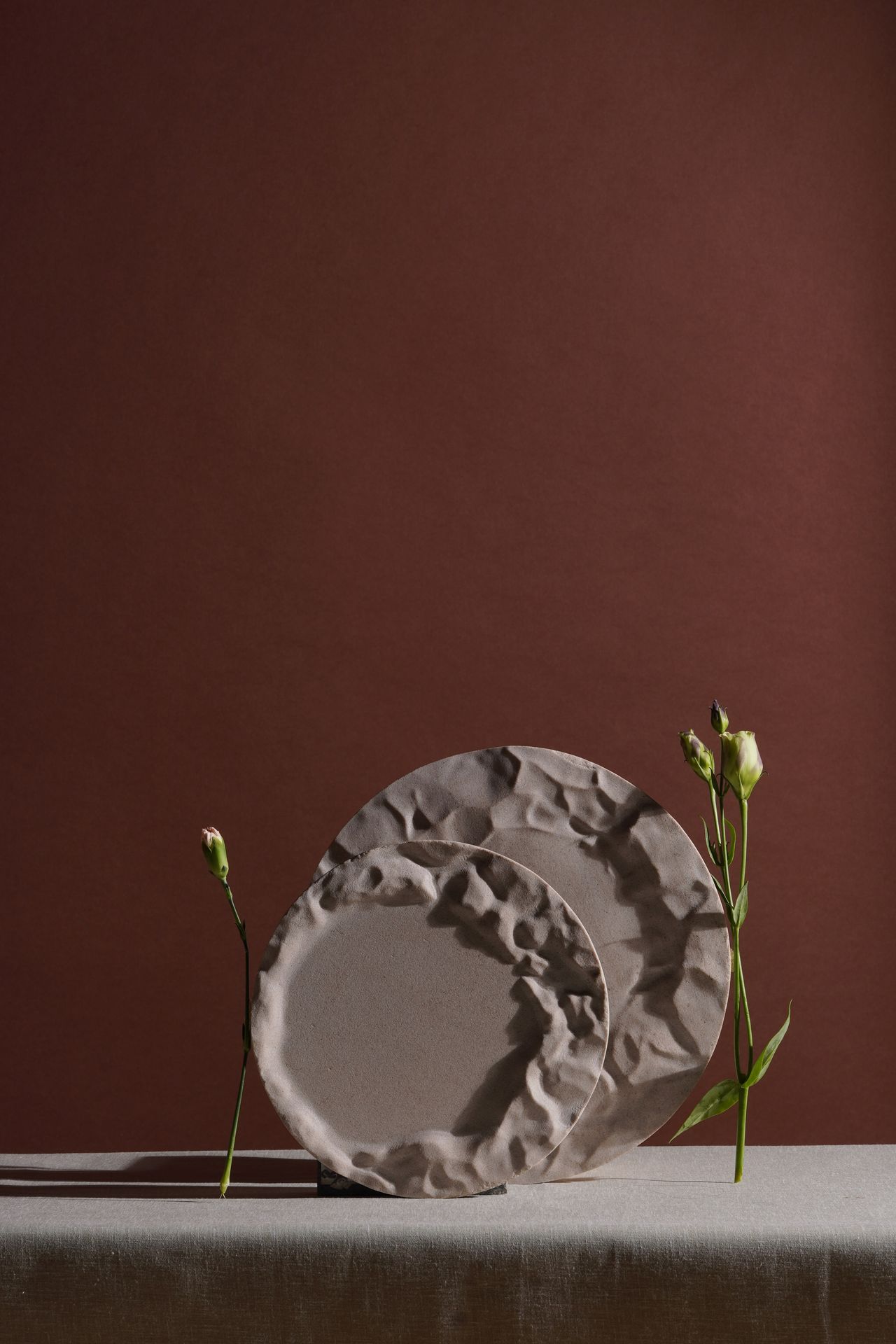 Two plates in grey stand vertically on a grey surface against a dark red background. Next to them are two white flowers, enclosing them on each side.