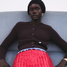 Model slouching on a grey sofa wearing a dark brown cardigan and red textured skirt sold at Selfridges