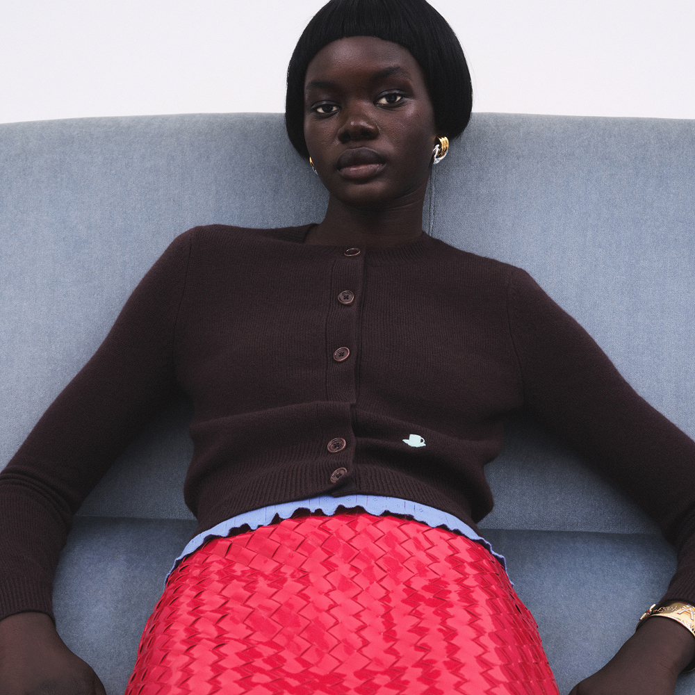 Model slouching on a grey sofa wearing a dark brown cardigan and red textured skirt sold at Selfridges