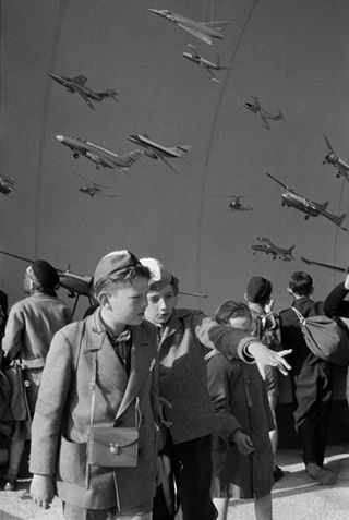 Black and white 1958 World's Fair in Brussels Image, two young boys dressed in caps and overcoats, looking at their surroundings, one boy is pointing, backdrop of multiple army planes flying in the sky
