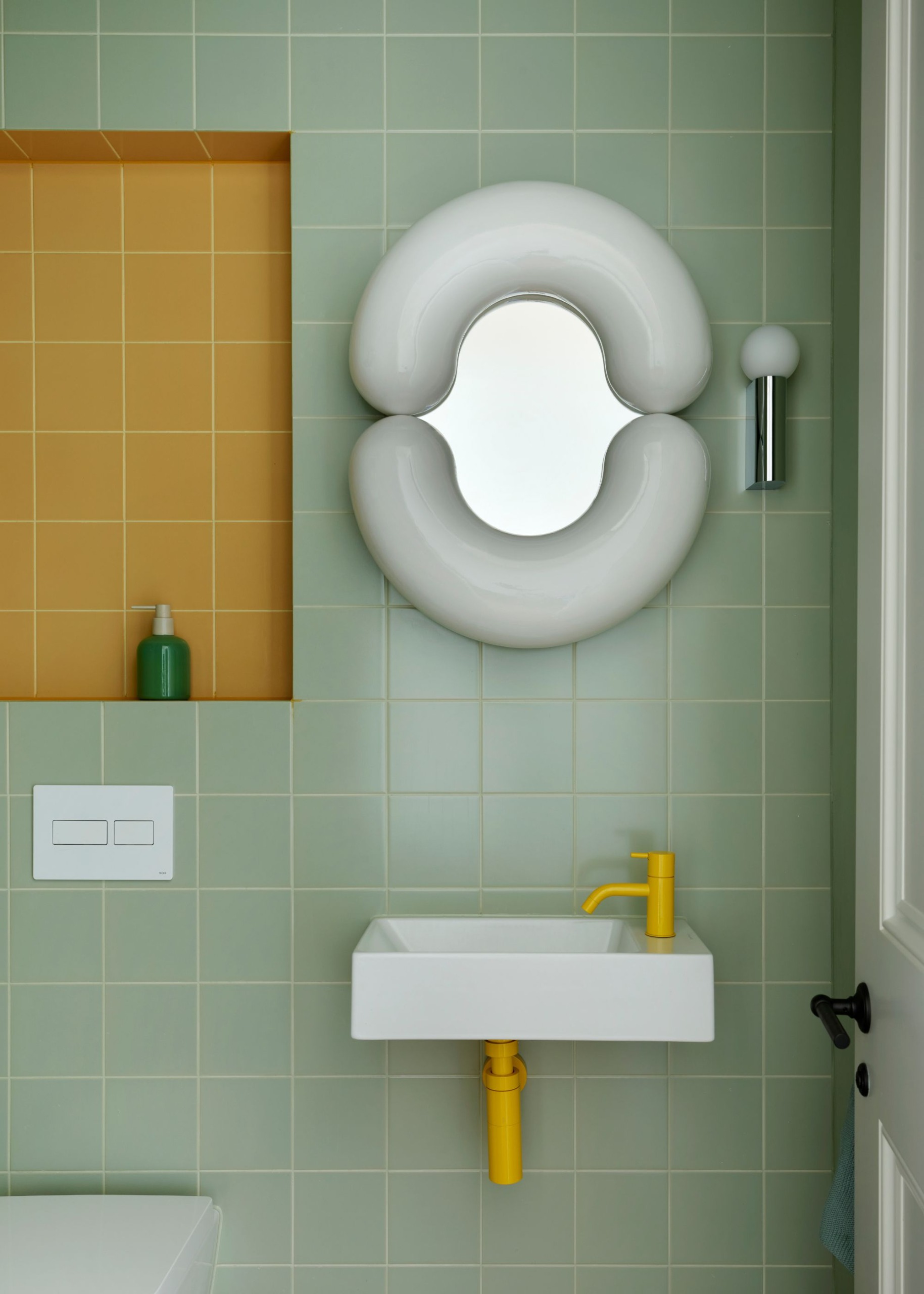 A small bathroom with square mint green wall tiles, a shelf recess with mustard yellow tiles, a small white sink with bright yellow hardware, and a modern, sculptural white mirror on the wall.