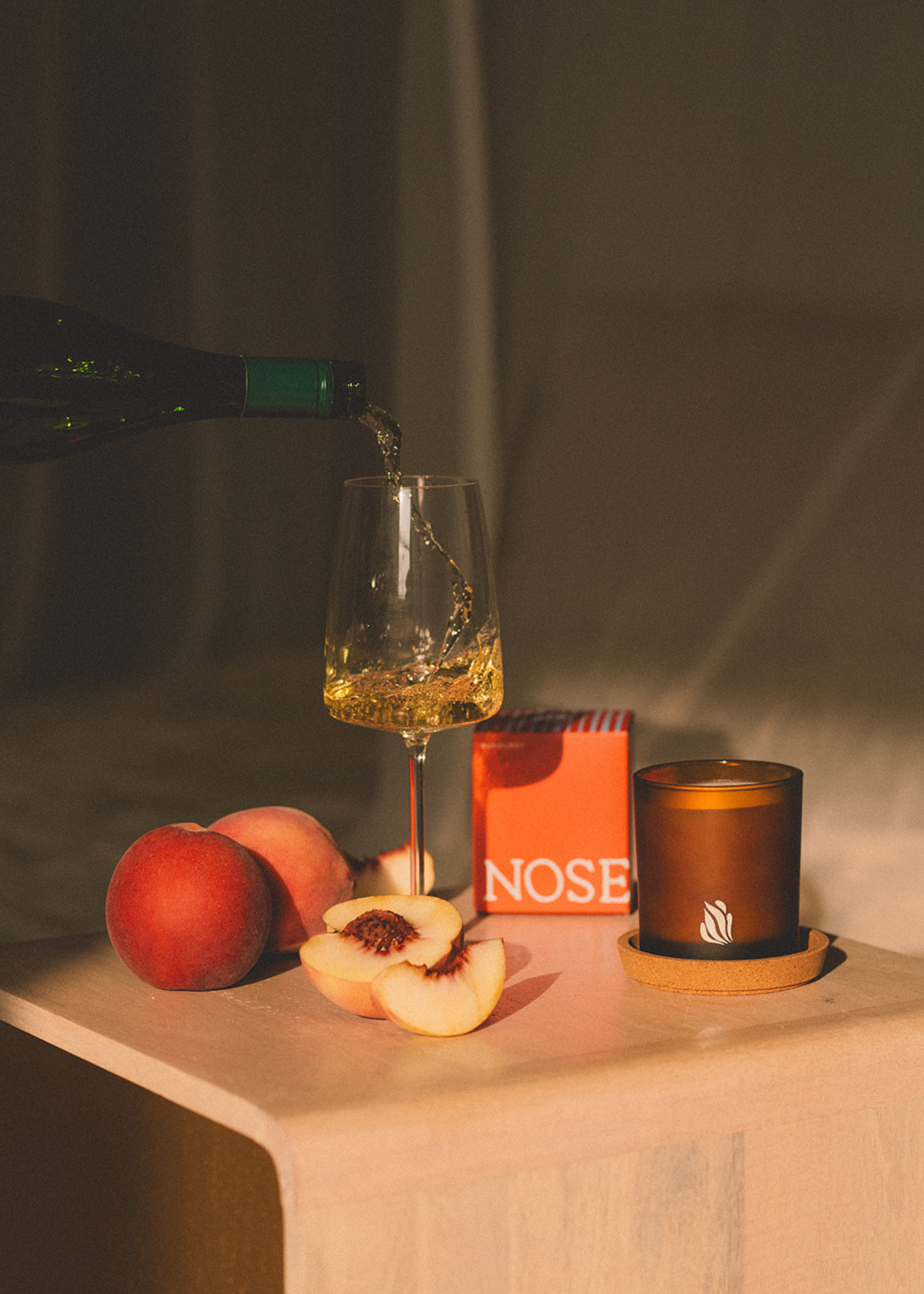 A candle from Nose Dive on a wood side table beside cut and whole peaches and a glass of white wine