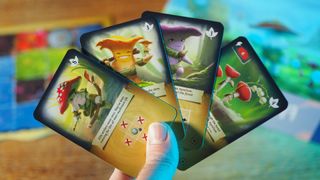 A hand holds up cards showing mushroom people from Mycelia, all against a wooden background with a board and the game box