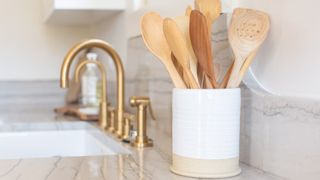 wooden utensils in a pot on a modern kitchen worktop, with a white sink and gold tap in the background
