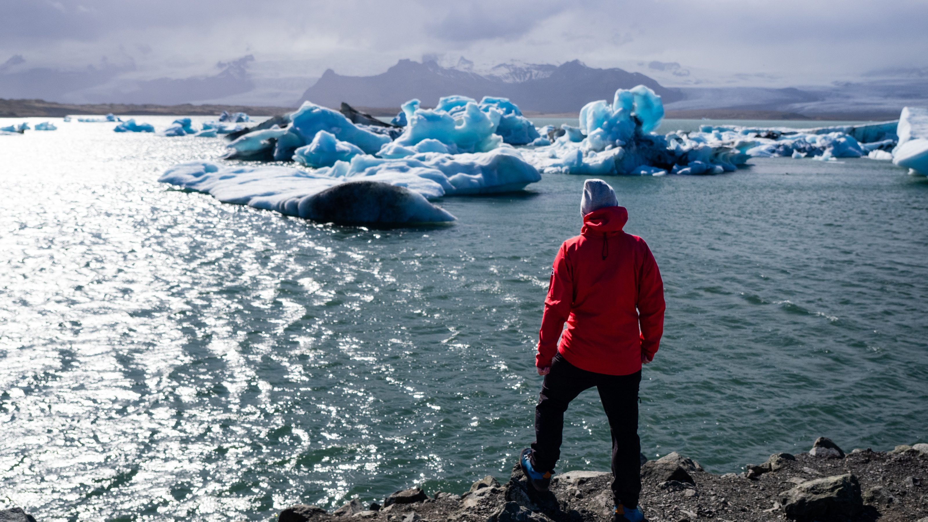 A man in a red jacket overlooks a serene body of water surrounded by small icebergs.