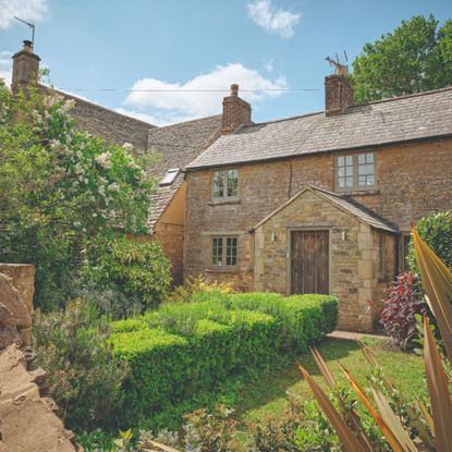 Exterior of bricked cottage house with lots of greenery in the front garden and a wooden door