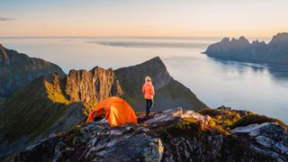 Woman next by a tent on mountain top contemplating the panorama