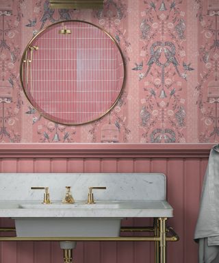 A small pink bathroom with blue and white bird wallpaper, a gold circular mirror, and a marble standing basin with a gold faucet and legs