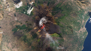 An aerial view of a volcano eruption. Slight bits of bright red lava are seen coming from the top of the volcano.