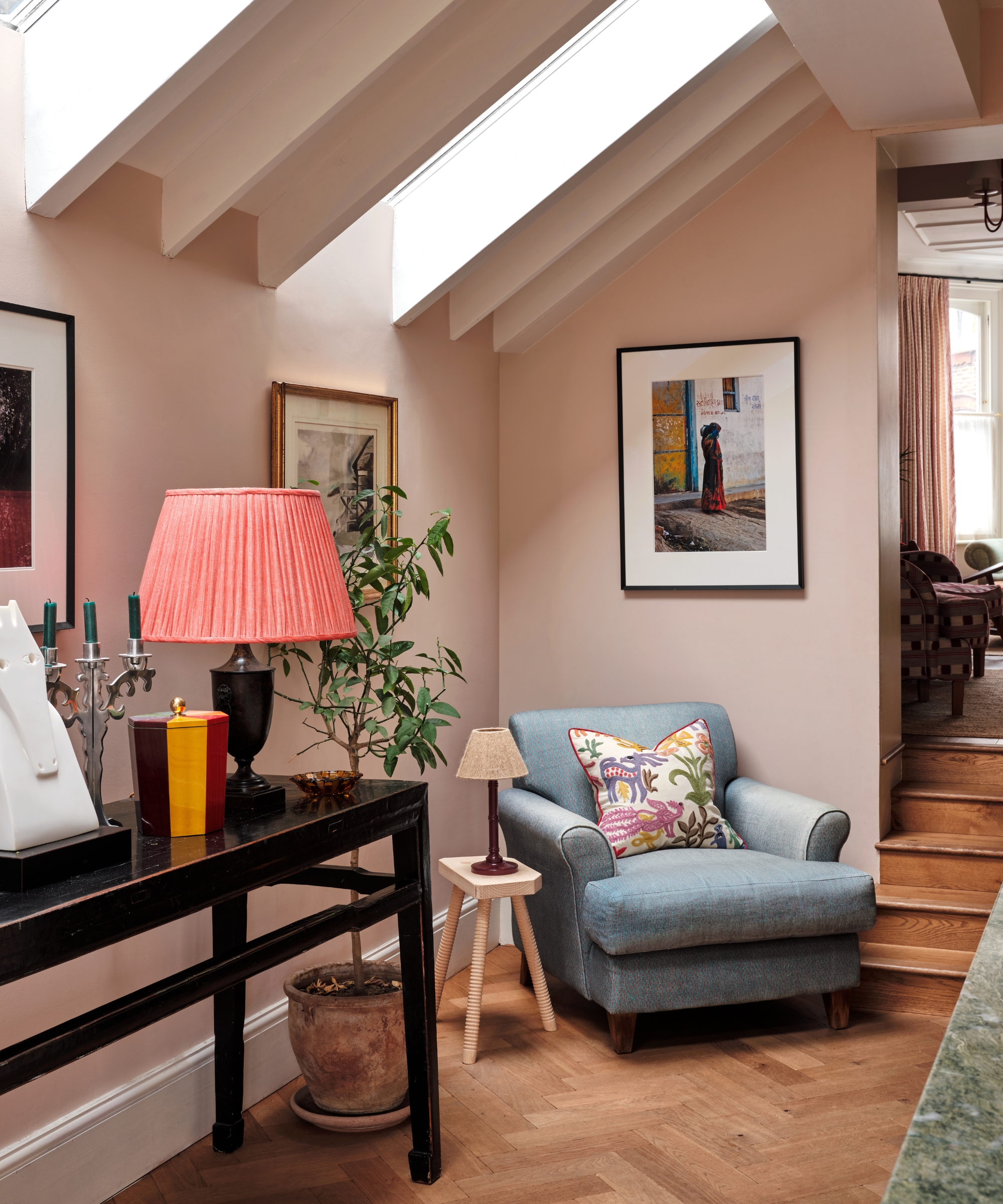 The corner of a kitchen with pink walls and chevron wood floors, featuring a blue armchair, and credenza table, and framed art on the walls