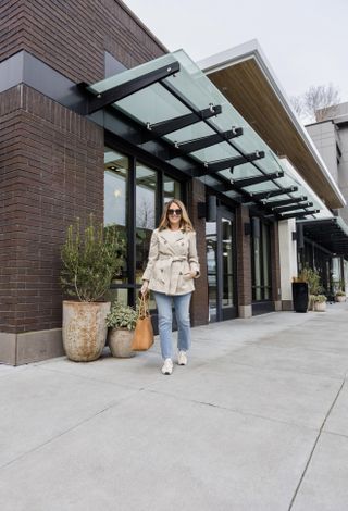 A Nordstrom stylist wearing a tan coat, jeans, sneakers, and sunglasses.