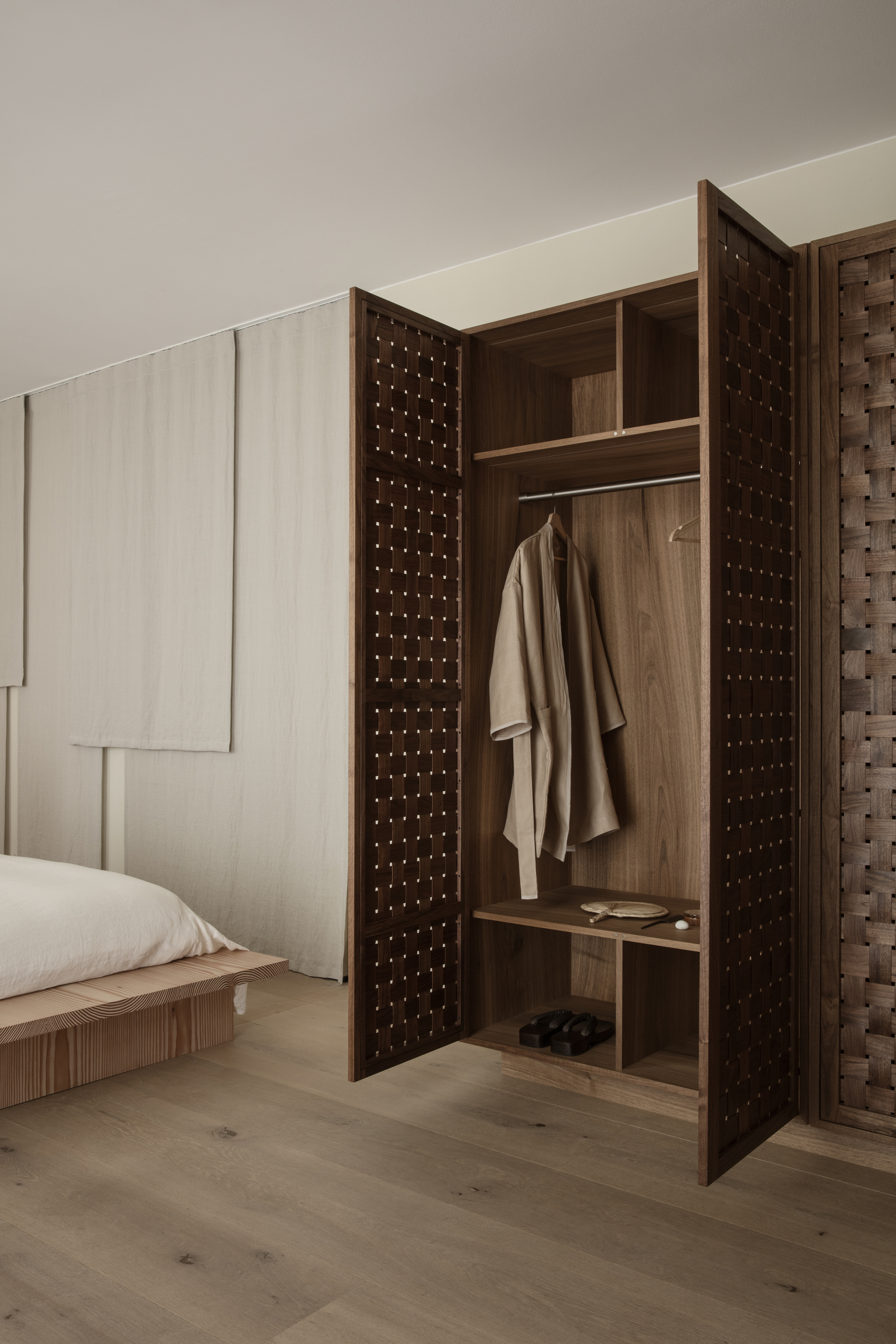 Image of a minimalist bedroom with a large, wooden built-in closet that is open and has a robe hanging in it.