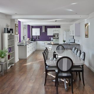kitchen area with white and purple wall and wooden floor and white kitchen units and dining table with chairs